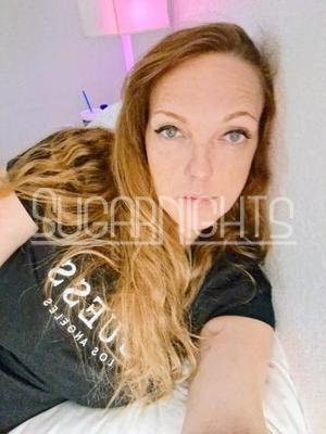 Elle wears a black logo t-shirt while resting on a bed. Elle wears a black logo t-shirt while resting on a bed.